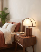 Halvo walnut nightstand lamp with a white half-dome shade lit on a wooden nightstand beside a bed