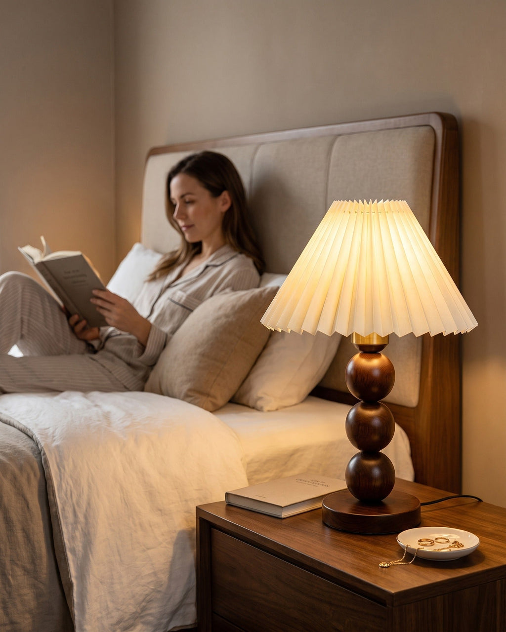 Bobbin Trio Wood Nightstand Lamp lit on a bedside table while a woman reads in bed