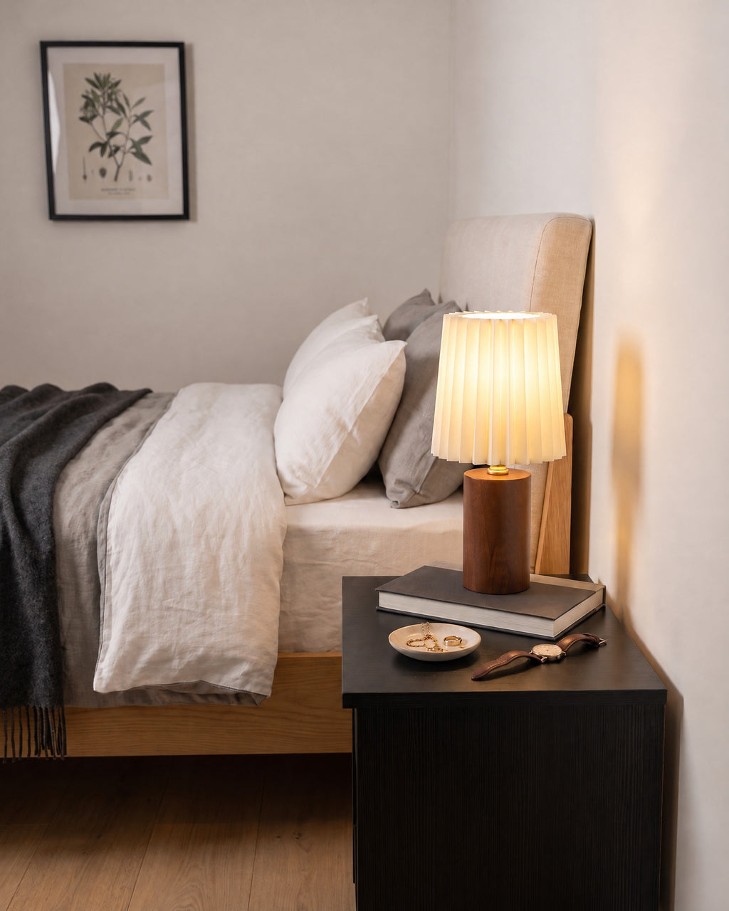 Linnea pleated walnut lamp on a black nightstand beside a bed, seen from the right