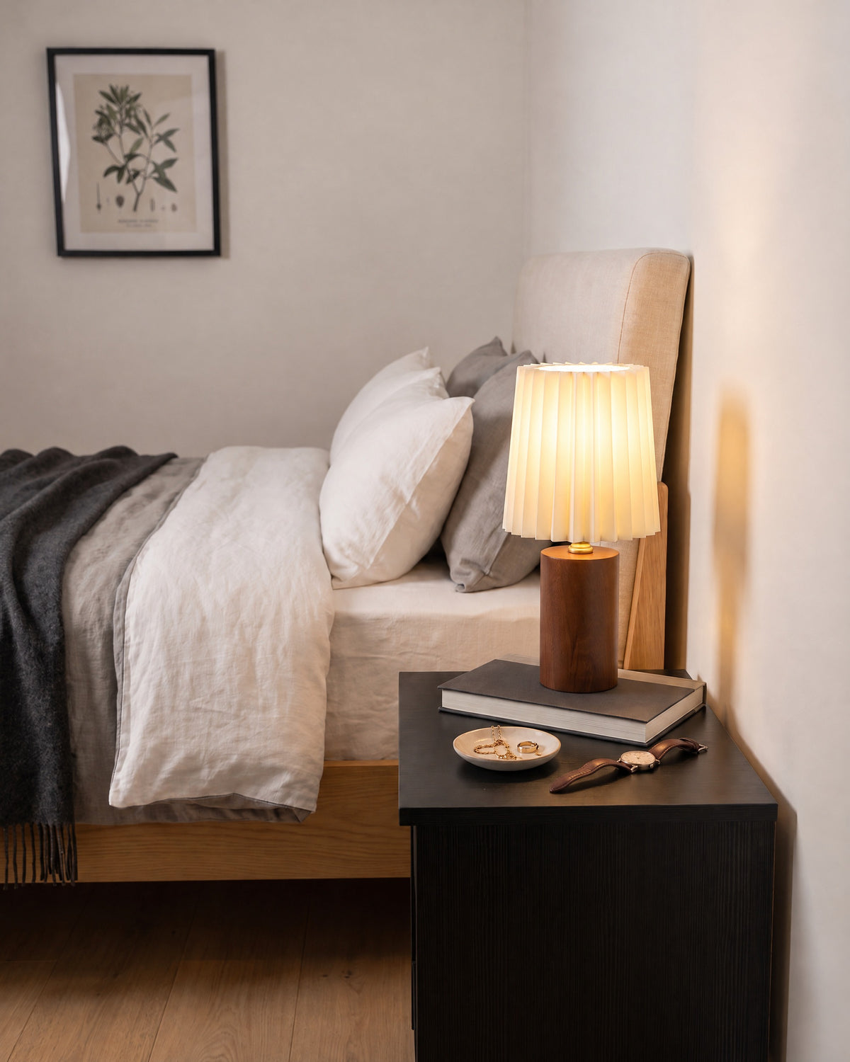 Linnea pleated walnut lamp on a black nightstand beside a bed, seen from the right