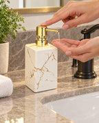 White: A hand pressing the pump of the Marble Soap Dispenser, Square Ceramic, Gold Pump